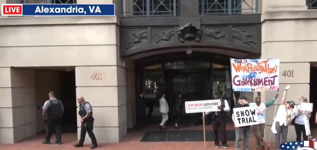 Protesters gather outside a government building in Alexandria, VA, holding signs addressing issues of government accountability and social unity.