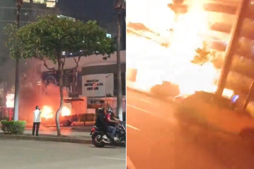 Fire erupts in an urban area, with onlookers observing the blaze near buildings and vehicles at night.