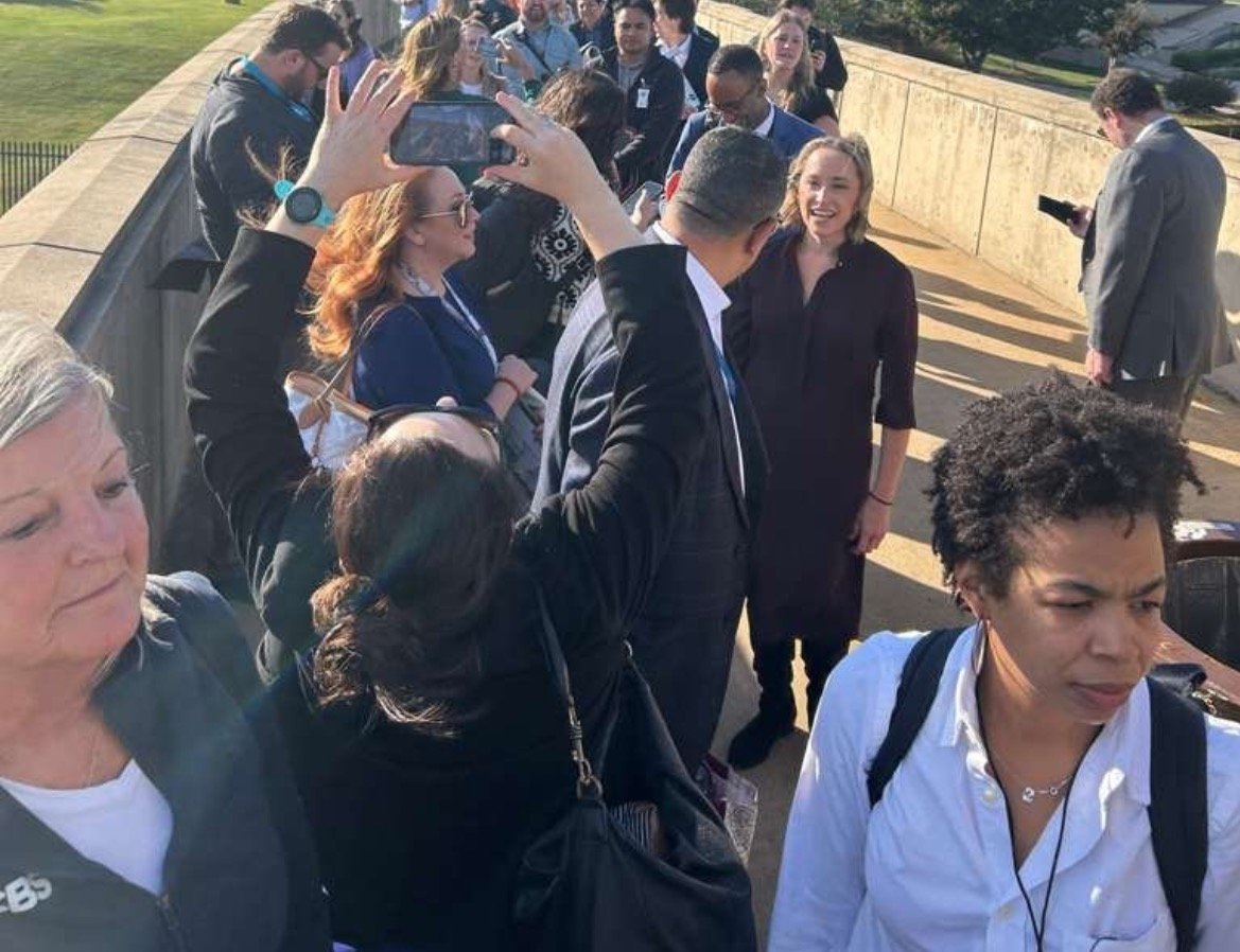 A diverse group of people gathered outdoors, with some taking photos and others engaged in conversation during a sunny day event.