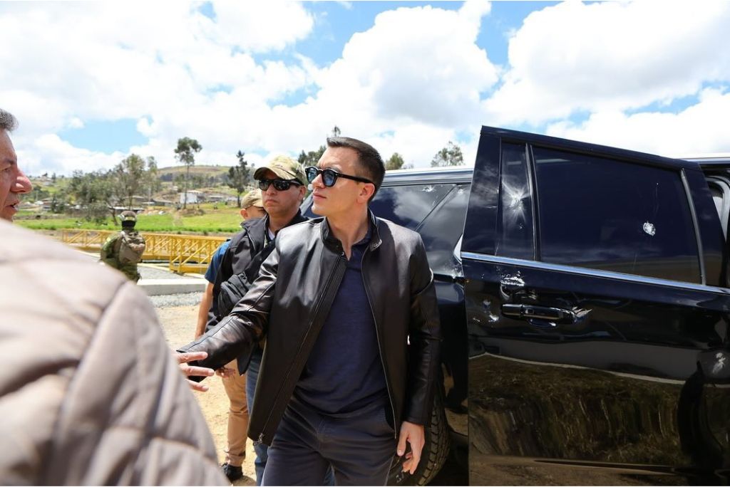 A man in sunglasses and a leather jacket engages in conversation near a black SUV, surrounded by onlookers and security personnel in a rural setting.