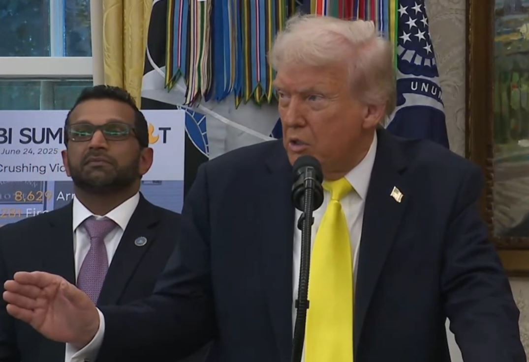 Donald Trump speaking at a podium in the Oval Office, with an official in a suit standing beside him, discussing the upcoming FBI Summit.